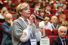 A person standing between rows of red auditorium seats asks a question during a conference, holding a microphone, surrounded by a large audience