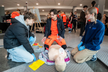 Three people participating in first aid training, kneeling around a resuscitation manikin and discussing the next steps; medical accessories used during the training are visible nearby.