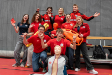 A group of women and men in red and navy rescue uniforms is posing together in a sports hall. Some of them are holding rescue equipment. They are all standing or kneeling against a sports net backdrop, creating a dynamic, cheerful group pose.