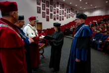 The graduate in an academic gown stands opposite four people and holds the document with both hands. She hands it to the professor in a light‑coloured gown, who reaches out to take it.