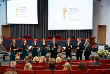 A vocal ensemble performs a piece during the ceremony in the auditorium of the Faculty of Pharmacy at the Medical University of Warsaw, standing before the audience against the background of two screens displaying the university’s logo