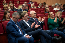 A few people are sitting in the front rows of red chairs and listening to a speech. Closest to the camera sit two men and two women, who are resting their hands on their laps or clasping them in front of themselves. In the further rows, a larger group of participants is taking their seats.