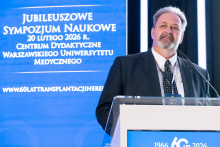 A person stands at the lectern, delivering a presentation during the symposium. They rest their hands on the podium and look toward the audience. Behind the speaker is a tall blue screen displaying information about the jubilee event.