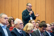 A person standing between rows of red auditorium seats asks a question during a conference, holding a microphone, surrounded by a large audience