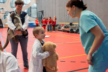 A boy wearing a white lab coat is holding a plush teddy bear and talking to a woman dressed in medical attire. Next to them stands a person with a large plush toy.