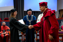 The graduate in an academic gown shakes hands with a professor in a red gown and looks in his direction. She holds a document in her other hand. Behind her stand three other people in ceremonial gowns.