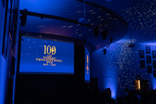 “A hall illuminated with blue lights during the ceremony; the screen displays the inscription ‘100 years of the Faculty of Pharmacy, WUM 1926–2026’, and light reflections are visible on the walls.