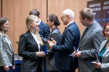 A group of several people is standing and talking after the award ceremony. Some of them are holding closed or open medal cases. The individuals are arranged in a loose line and are turned toward one another, gesturing as they talk. In the background, wooden panels and part of the event’s roll‑ups can be seen.