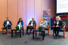 A discussion panel with five people seated on stage in armchairs; one person holds a microphone and moderates the conversation. Tables with water bottles stand in the center, and a presentation screen and conference roll‑up are visible in the background