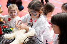 A girl wearing a white lab coat is examining a plush teddy bear, holding a plastic syringe to it. Next to her, other children participating in a medical-themed activity are sitting at the table in the sports hall.