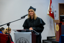 A woman in an academic gown stands behind the lectern and addresses the participants of the ceremony. She looks straight ahead and keeps her hands folded on the lectern. The microphone is positioned right next to her face.