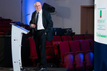 A speaker stands at the lectern, delivering a presentation. He looks toward the large screen, which displays a quote about sharing knowledge. He holds a clicker in one hand and rests the other on the podium. The screen’s background features green‑and‑blue hexagonal patterns.
