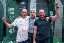 Three men are standing next to each other and raising their clenched fists in a gesture of joy. Two of them have their arms around the person in the middle. They are all looking toward the camera and appear to be smiling. Behind them are green flags with the logo of a sports organization.