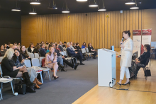 Sala konferencyjna z drewnianymi ścianami i ciemnym sufitem. Po lewej stronie siedzi kilkanaście osób na krzesłach. Po prawej stronie osoba prowadząca stoi przy białej mównicy i mówi do publiczności. Za prowadzącą znajduje się duży ekran z jasnym slajdem prezentacji.