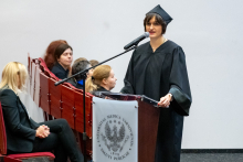 A woman in an academic gown stands by the lectern and addresses the audience. She rests one hand on the lectern and slightly raises the other. She is looking straight ahead.