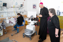A clergyman dressed in black is standing next to a woman while visiting a room with newborns. In front of them stands a cart with medical equipment. On the left, a woman is sitting on a chair beside an incubator and leaning over a baby. In the background, monitors and other room equipment are visible.