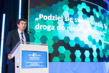 A person standing at a lectern during a scientific conference, against the backdrop of a large screen displaying the quote: ‘Share your knowledge. It is the path to immortality.’ Next to it, there is a sign informing about the anniversary symposium on kidney transplantation in Poland