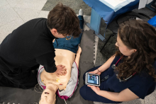 A person performing chest compressions on a manikin during cardiopulmonary resuscitation training, while another person nearby monitors the parameters on the training device.