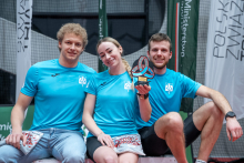 Three people are sitting next to one another and smiling. The person in the middle is holding a sports trophy raised in the air. The other two are leaning slightly toward each other and holding small gifts in their hands. They all appear happy and are celebrating a shared success. Behind them, vertical banners of sports organizations are visible.”