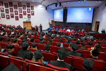 Many people in academic gowns are seated in rows, looking toward the lectern. In front of them stands the graduate in an academic gown, delivering a speech.