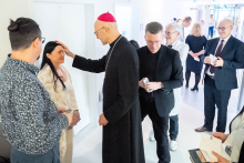 A person dressed in a long, dark outfit with a red head covering is standing in a bright corridor and making a gesture toward another person. Around them, several people are standing in a queue and others are walking further down the corridor.