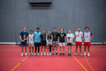 Ten people are standing in a single row in a sports hall. Several of them are holding trophies, while the others have medals around their necks. They are all looking toward the camera and appear pleased. The group is presenting the awards they received after the competition.