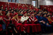 University authorities in ceremonial gowns are seated in the front row, applauding. Next to them sit the graduates in black gowns. Behind them, additional rows of participants observe the course of the ceremony. Everyone is looking toward the stage.