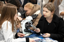 People talking at a laboratory station, where a microscope and informational materials are placed on the table; one of the individuals is holding a leaflet while discussing the presented topics.