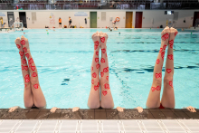 Three people are lifting their legs vertically above the water at the edge of a swimming pool. Each pair of legs is covered with numerous red WOŚP heart stickers. The people are supporting themselves with their hands on the pool edge. In the background, other participants are swimming and taking part in the activities.