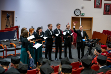 Seven people stand in a semicircle and perform a piece during the ceremony. They are holding open songbooks and looking toward the audience. One person raises a hand and conducts. In the front rows, the participants of the ceremony are seated.