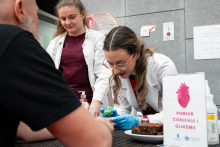 Two female students in medical coats are measuring the blood glucose level of a man sitting at a small table. One of them is holding his hand and using a small device, while the other is observing the examination. Informational materials and snacks are on the table.