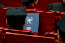 The graduate holds a folder with the university’s logo and rests it against the back of a red chair. He is seated among other participants of the ceremony, who are also wearing gowns.
