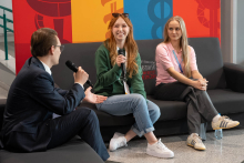 Three people sitting on a couch during a discussion at the event; two of them are holding microphones, leading the conversation against a colorful, graphic wall.