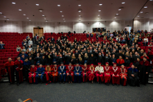 A large group of graduates and university staff pose together in the auditorium. In the front row, the university authorities in ceremonial gowns sit and look toward the photographer. Behind them stand dozens of graduates in gowns, smiling, waving, and raising their hands in a gesture of joy. In the background, additional participants are seated.