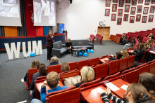 A person delivering a presentation on stage, holding a microphone and a slide clicker, standing in front of large screens displaying educational illustrations and next to decorative WUM letters.