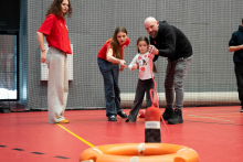 Five people are participating in a rescue exercise in a sports hall. Two adults are helping a child throw a rope toward a lifebuoy lying on the floor. A woman next to them is pointing out the direction and giving additional instructions. Another person is standing aside and observing the task.