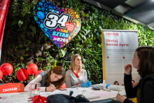 Three women are sitting at a table during a charity event. One of them is filling out a form, another is providing information, and the third is talking to someone outside the frame. Documents and DKMS materials are lying on the table. In the background, there is a large colorful heart with the logo of the 34th Grand Finale of the Great Orchestra of Christmas Charity, as well as balloons and a campaign roll‑up.