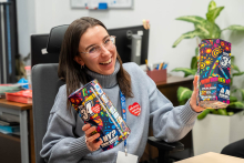 One woman is sitting at a desk and holding colorful fundraising tins in both hands. She is smiling and presenting them in front of her. A red WOŚP heart is visible on her sweater.
