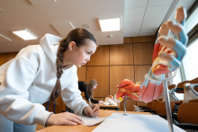 A person leaning over a table in a teaching room, taking notes, with an anatomical model of a limb’s musculature beside them; in the background, another person is working at a different station.