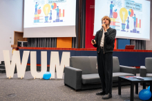 A person delivering a presentation on stage, holding a microphone and a slide clicker, standing in front of large screens displaying educational illustrations and decorative “WUM” letters placed on the floor.