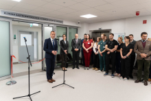 A group of medical staff and officials standing together during an opening event in a hospital corridor, with a speaker addressing the audience near a microphone stand.