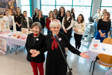 Osoba stojąca przy mikrofonie przemawia podczas wydarzenia edukacyjnego. Obok stoi druga osoba, a za nimi ustawiona jest grupa uczestników. W tle widoczne są stoliki z materiałami informacyjnymi i planszami edukacyjnymi.