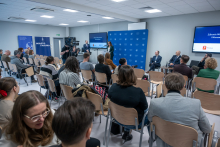 Around twenty people are sitting in a room and listening to a presentation. Three speakers are seated at the front, and one person is standing at the lectern, addressing the audience. Several people are operating a camera positioned on the left. The participants are looking toward a screen displaying content related to the health of Warsaw’s residents. The atmosphere in the room is calm.