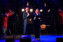 Two people are standing next to each other on stage, hosting the event. The man is holding a microphone and speaking. The woman is holding papers and looking toward the audience. Both are dressed elegantly. Behind them, bright spotlights are shining, and the background is filled with blue light dots.