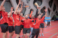The team in red jerseys is celebrating their victory in the sports hall, lifting the trophy and raising their hands in joy, while other participants watch them from the side of the court.