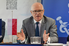 A middle‑aged man wearing glasses — the rector of WUM — is sitting at a table and gesturing with his hands. He is dressed in a grey suit and a blue tie.