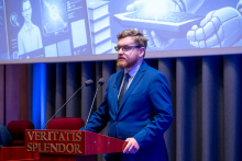 A person in a navy-blue suit is standing at a lectern with the inscription ‘Veritatis Splendor’, speaking into two microphones. In the background, a large screen displays a futuristic graphic of a robot and a technological interface.