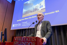 A person standing at a podium during a presentation, with a large screen behind them displaying a slide featuring information and an image of UMass Chan Medical School buildings in Worcester.