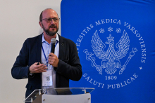 A middle‑aged man, dressed formally and wearing glasses, is standing behind a lectern. He is holding a microphone in his left hand and speaking into it. Behind him is a blue banner with the white WUM logo — an eagle and stars surrounded by the university’s Latin name.