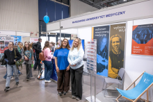 In front of the Medical University of Warsaw’s booth, two people are standing and posing for a photo. Posters about study programs are displayed at the booth, and next to it there is a stand with leaflets. In the background, participants of the fair can be seen walking by
